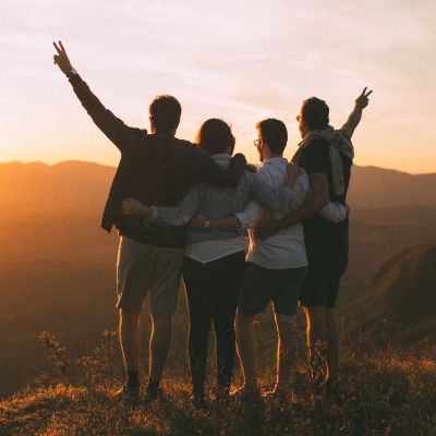 Gruppe mit vier Personen, die ihren erfolgreichen Bergaufstieg auf dem Gipfel genießen. Rechts und links außen haben die beiden Personen den Arm jeweils in die Luft gereckt. Alle stehen mit dem Rücken zum Betrachter und schauen in den Sonnenuntergang.