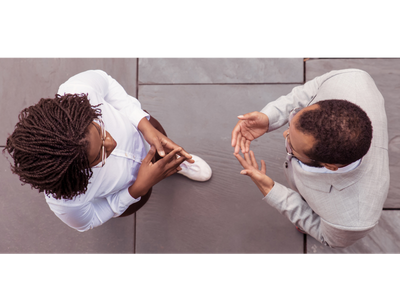 Zwei Personen im Gespräch von oben betrachtet, einander zugewandt und gegenüberstehend. Beide in Bürokleidung, weißes Hemd links, hellgraues Jacket und weißes Hemd rechts. Beide mit dunkler Hautfarbe, Fliesenboden, auf dem sie stehen ist dunkelgrau