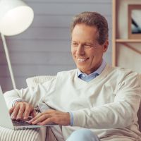 Mann im Sessel am Laptop Mann im Sessel am Laptop, lächelnd mit Blick auf das Display. Alles in Weiß, Beige, Hellbraun und Hellblau gehalten.