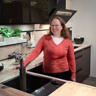 Frau steht lächelnd hinter dem Tresen mit der Spüle in der Küche, deren Fronten in Schwarz gehalten sind. Man sieht Küchenequipment.