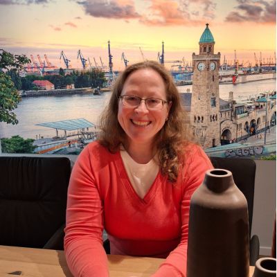 Frau sitzt am Tisch und lächelt in die Kamera, im Hintergrund ist eine von hinten beleuchtete Leinwand mit dem Motiv des Hamburger Hafens.