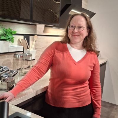 Frau steht lächelnd hinter dem Tresen mit der Spüle in der Küche, deren Fronten in Schwarz gehalten sind. Man sieht Küchenequipment.