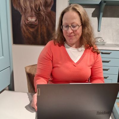 Melanie am Laptop Laptop auf Holztisch und Frau schaut lächelnd auf das Display vom Laptop, welches man nicht sehen kann. Ansicht auf Augenhöhe.