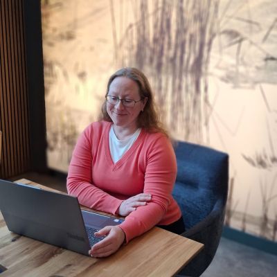melanie-laptop-2 Laptop auf Holztisch und Frau schaut lächelnd auf das Display vom Laptop, welches man nicht sehen kann. Ansicht leicht von oben